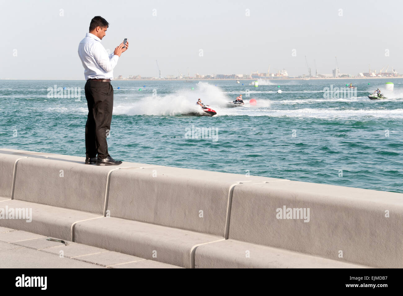 Un uomo si erge sul lungomare di prendere un telefono cellulare immagine un jet-ski la concorrenza a Doha, in Qatar. Foto Stock