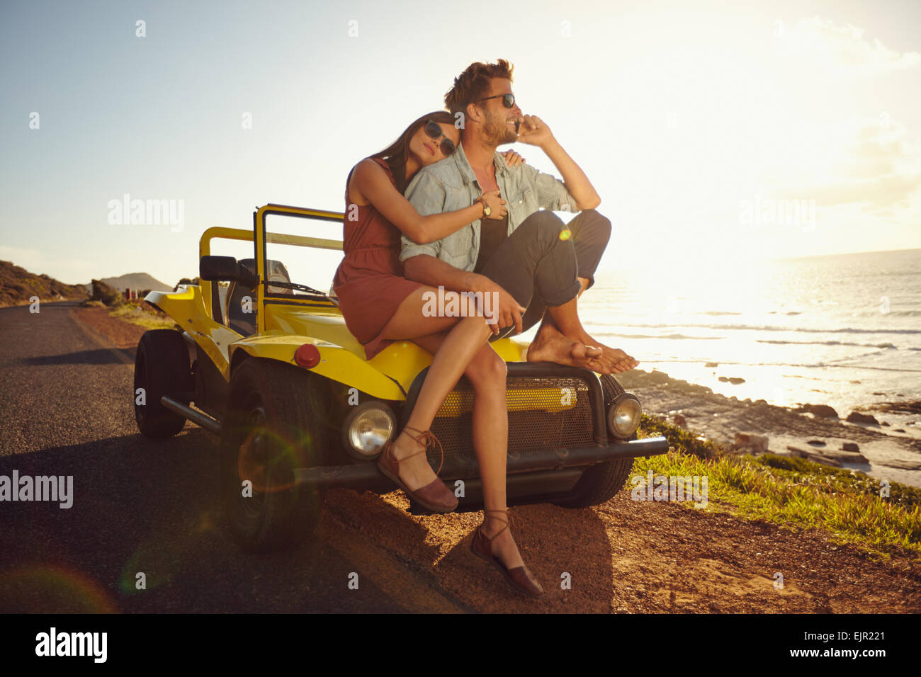 Una giovane coppia in viaggio. Coppia giovane seduto sul cofano della loro auto con l'uomo risponde a una telefonata e donna seduta da. Foto Stock