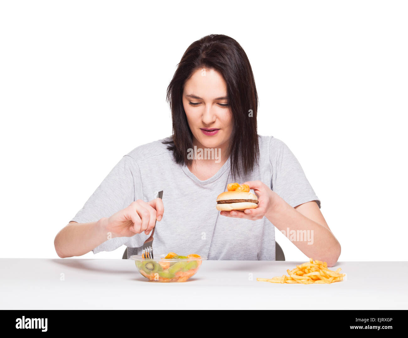 Una sana contro il cibo spazzatura concetto con un naturale donna ansante davanti pasto frutti e scegliendo le patatine fritte con hamburger, isolati o Foto Stock