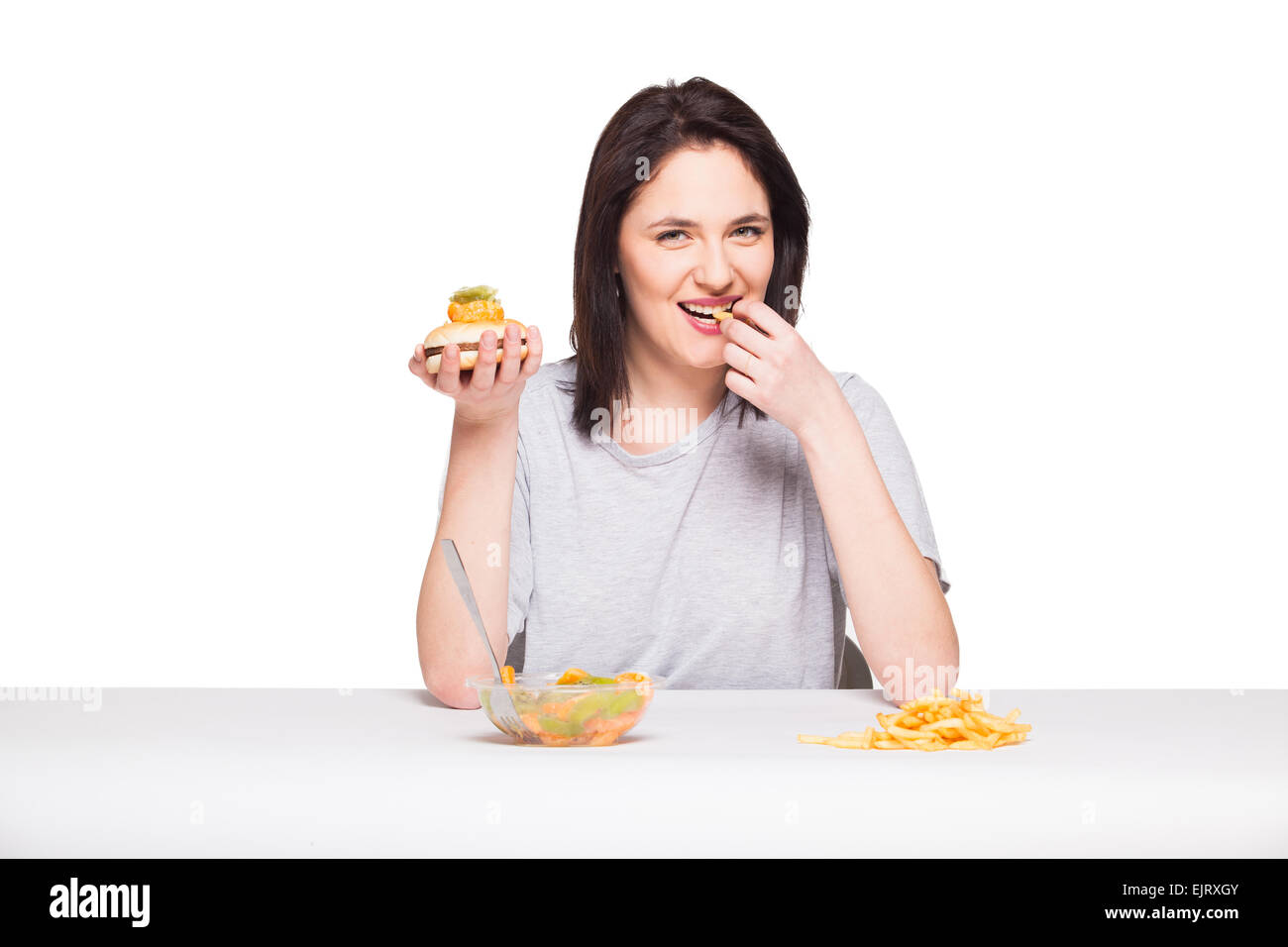 Una sana contro il cibo spazzatura concetto con un naturale donna ansante davanti pasto frutti e scegliendo le patatine fritte con hamburger, isolati o Foto Stock