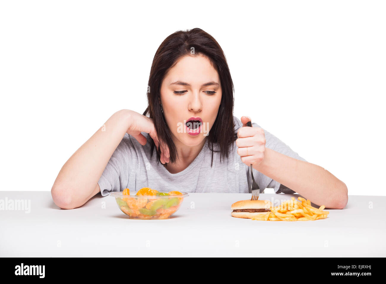 Una sana contro il cibo spazzatura concetto con un naturale donna ansante davanti pasto di frutta e patate fritte con hamburger, isolato su bianco Foto Stock