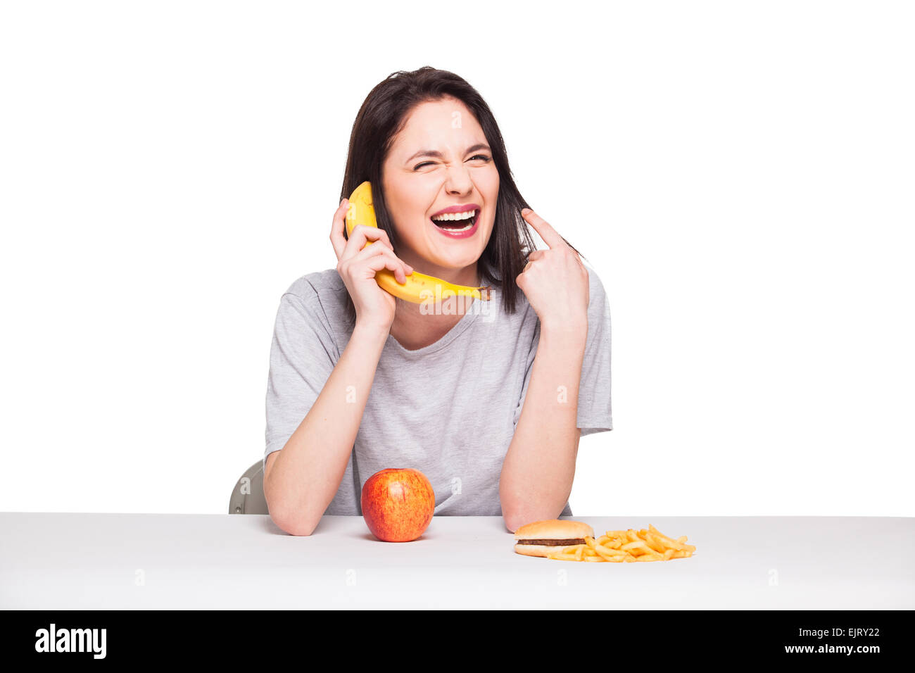 Naturale donna espressiva giocando con frutti, avendo di fronte junk e cibo sano, isolato su bianco Foto Stock