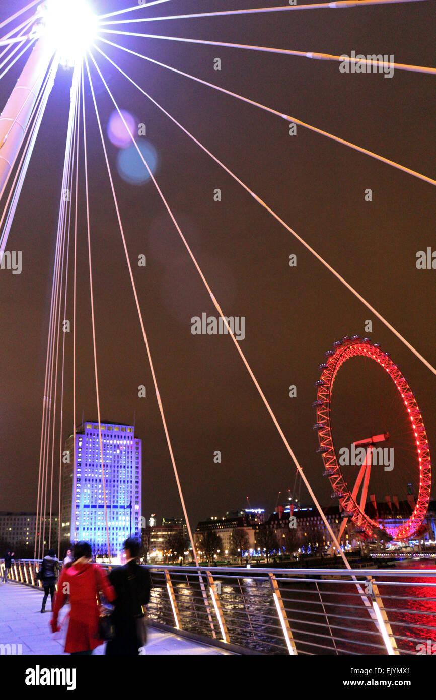 Londra England Regno Unito. Il 2 aprile 2015. Dopo il tramonto sulla Southbank London Eye è illuminato con le luci rosse della attuale sponsor Coca Cola e il guscio edificio si illumina in blu. Come si vede dal Giubileo ponte pedonale attraverso il fiume Tamigi. Foto Stock
