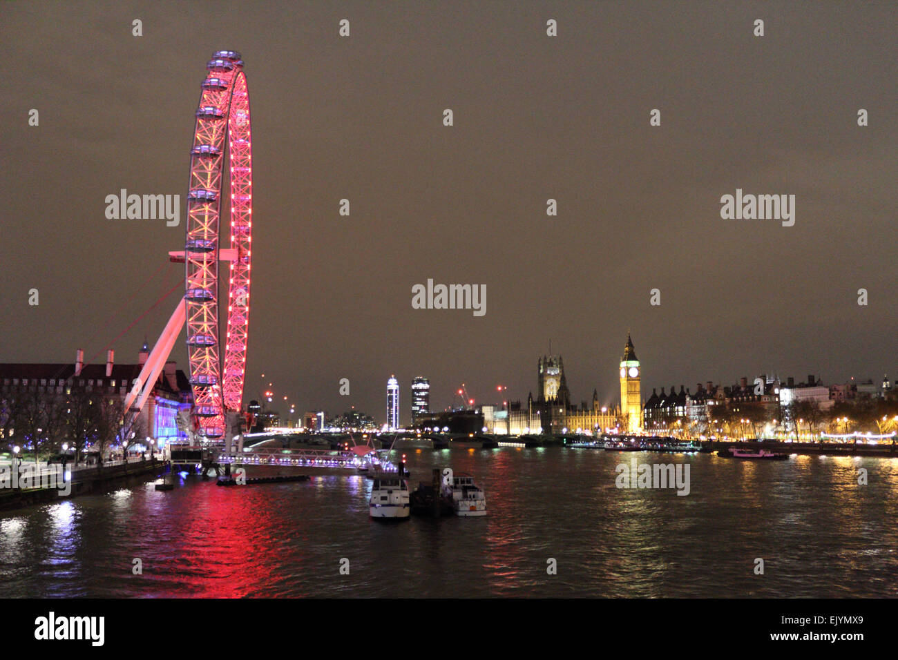 Londra England Regno Unito. Il 2 aprile 2015. Dopo il tramonto sulla Southbank London Eye è illuminato con le luci rosse della attuale sponsor Coca-Cola, con le case del Parlamento al di là. Come si vede dal Giubileo ponte pedonale attraverso il fiume Tamigi. Foto Stock
