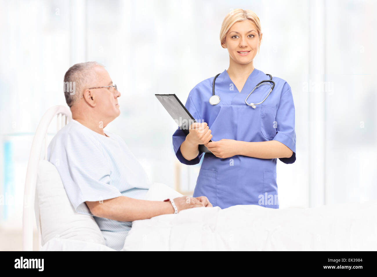 Femmina professionista sanitario tenendo un blocco appunti e in piedi accanto ad una matura paziente che giace nel letto di ospedale Foto Stock