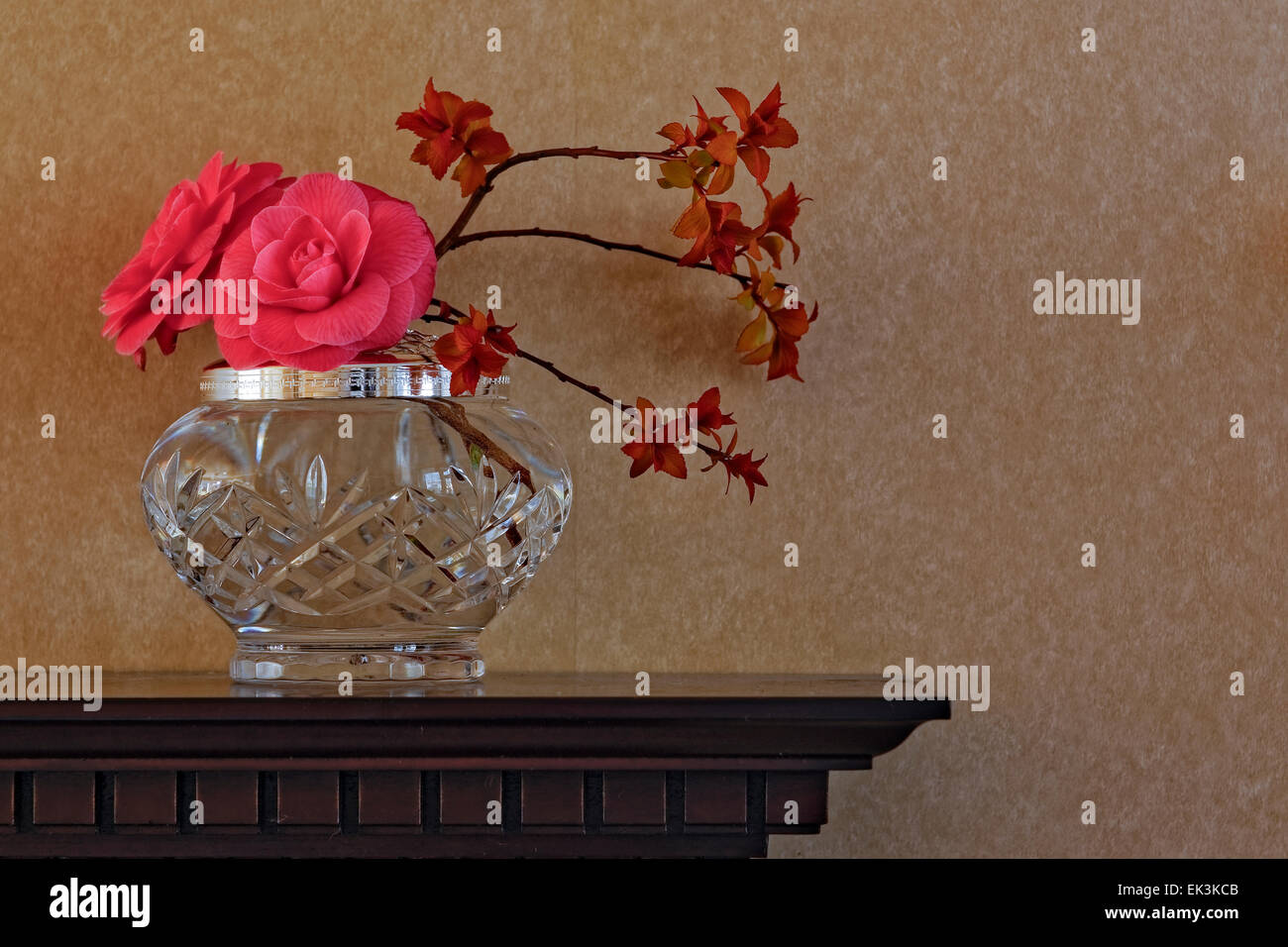 La composizione floreale, compresi camellia fiori in un vaso ​crystal su legno intagliato mantelpiece contro uno sfondo semplice; l'orientamento orizzontale. Foto Stock