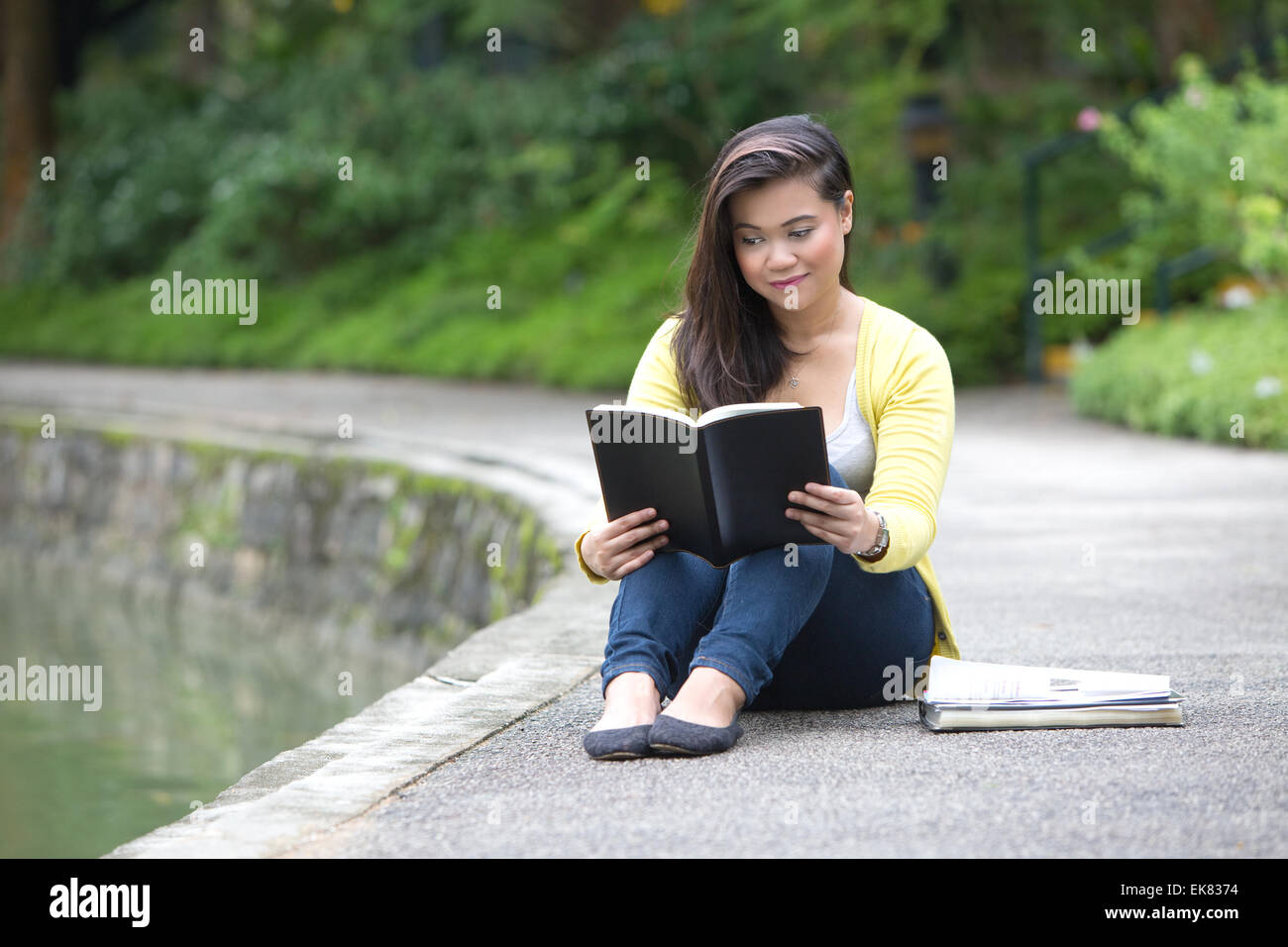 Bella giovane femmina università o college studente la lettura di un libro, seduto da un lago in un parco. Foto Stock