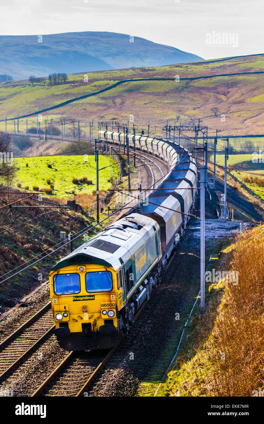 Treno per il trasporto di carbone a Cumbria, Regno Unito aprile 2015. Meteo Regno Unito. Giorno di sole luminoso sopra le campane di Cambrian ed il traffico ferroviario di West Coast Line del Freight. Un Freightliner 66560 Classe 66 trasporto container treno di carri a tramoggia di carbone alimenta a nord sulla West Coast Main Line, a Tebay. La maggior parte dei combustibili fossili estratti e degli aggregati vengono spostati in carri a tramoggia o gondole utilizzate per l'esportazione di carbone opencast estratto scozzese in centrali elettriche inglesi a carbone. Foto Stock