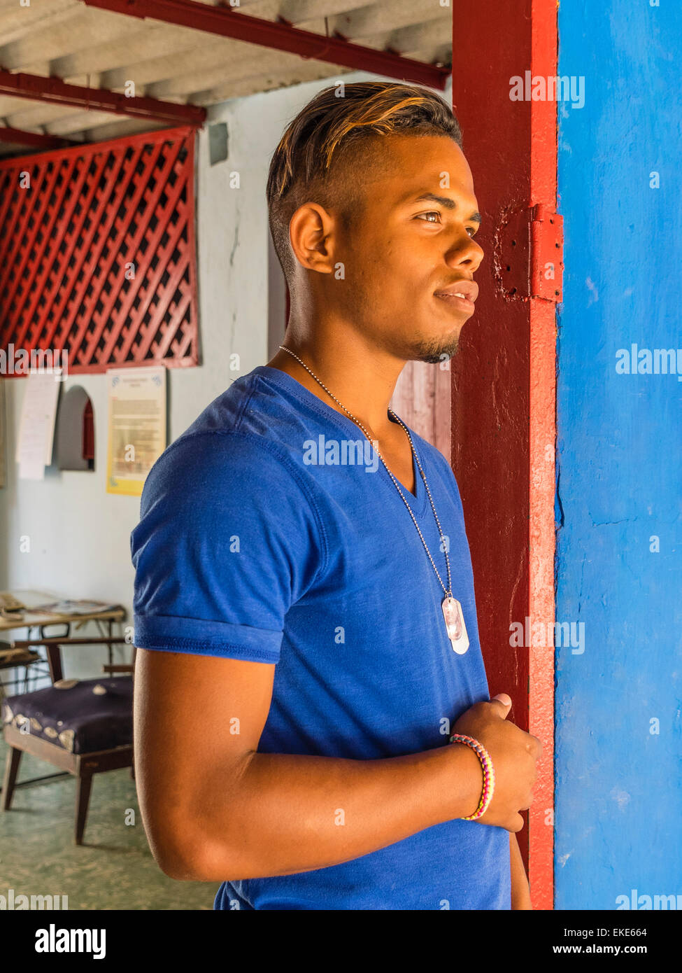Una ventina di qualcosa di afro-cubane l uomo sta in un molto colorato di rosso porta con una parete blu che corrisponde al colore della sua maglietta. Foto Stock