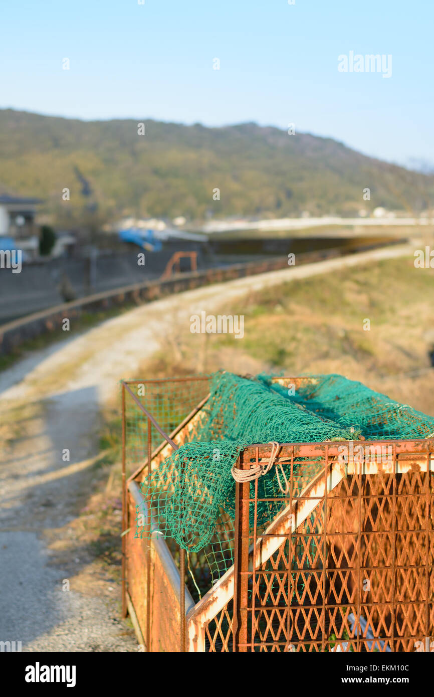 Un arrugginito contenitore di rifiuti nei pressi di un percorso sterrato seguendo la curva di un fiume e le montagne nella parte posteriore nella campagna giapponese. Foto Stock