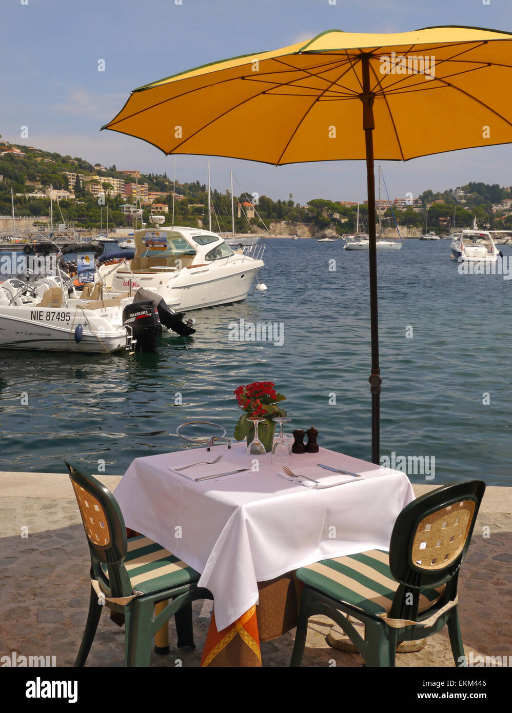 Un tavolo per la cena in un ambiente sul fronte mare in un porto con un ombrello giallo e barche in background. Foto Stock