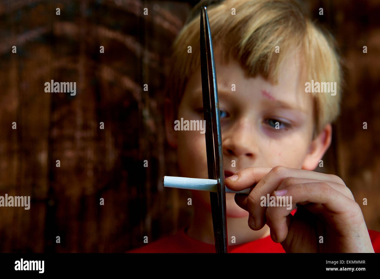 Close up bambino ragazzo biondo sigaretta di taglio con forbici e pareti in legno sfondo Foto Stock