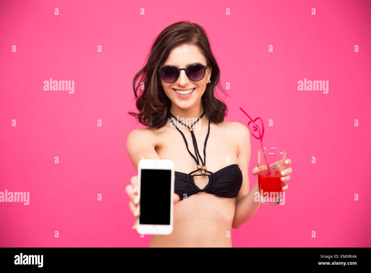 Donna sorridente in bikini e occhiali tenendo un bicchiere di succo di frutta e mostra lo schermo dello smartphone su sfondo rosa Foto Stock