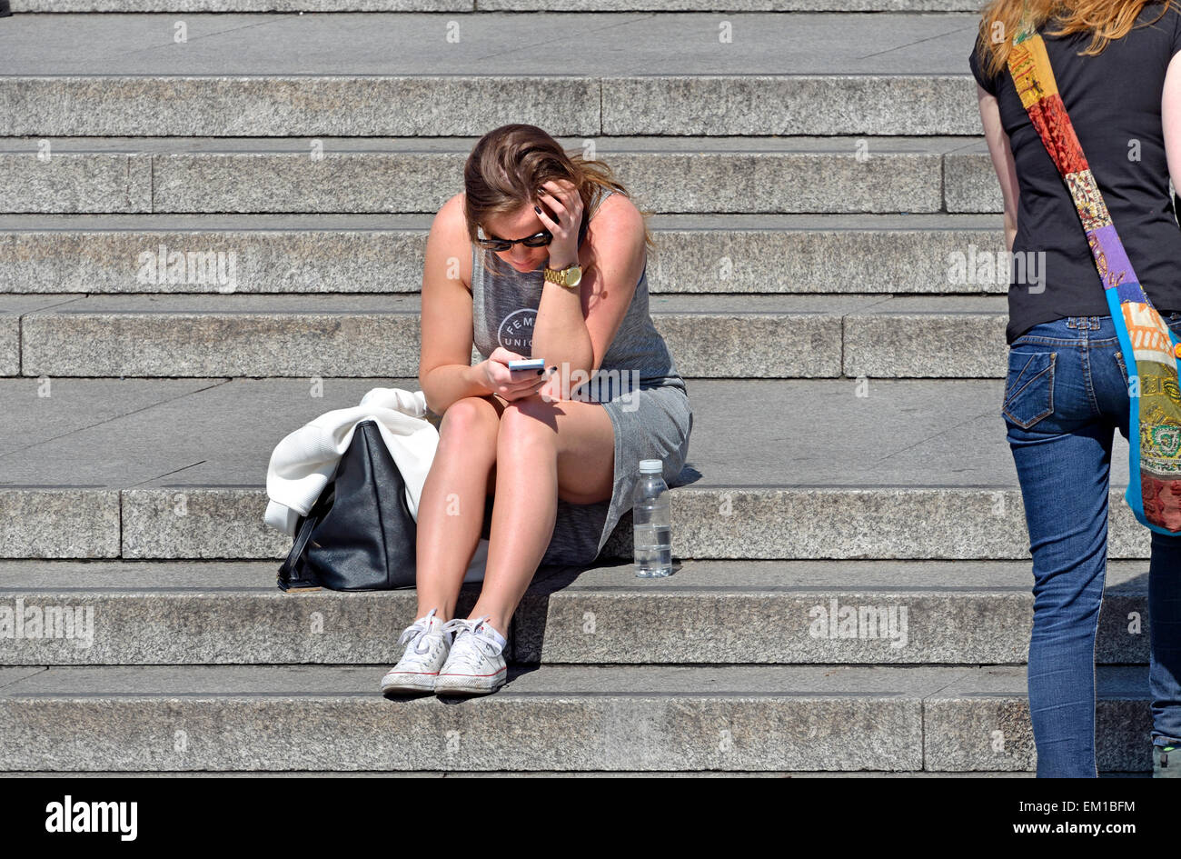 Londra, Inghilterra, Regno Unito. Giovane donna seduta sui gradini in Trafalgar Square, sms sul suo telefono cellulare Foto Stock