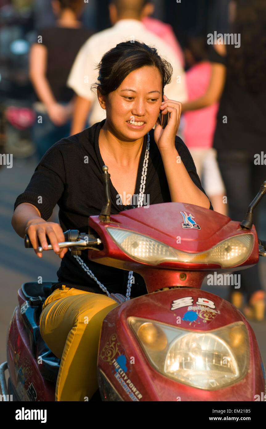 Donna Chat sul telefono mentre cavalcate schooter elettrico a casa dal lavoro, Lianyungang, Jiansu Provincia, Cina. Foto Stock