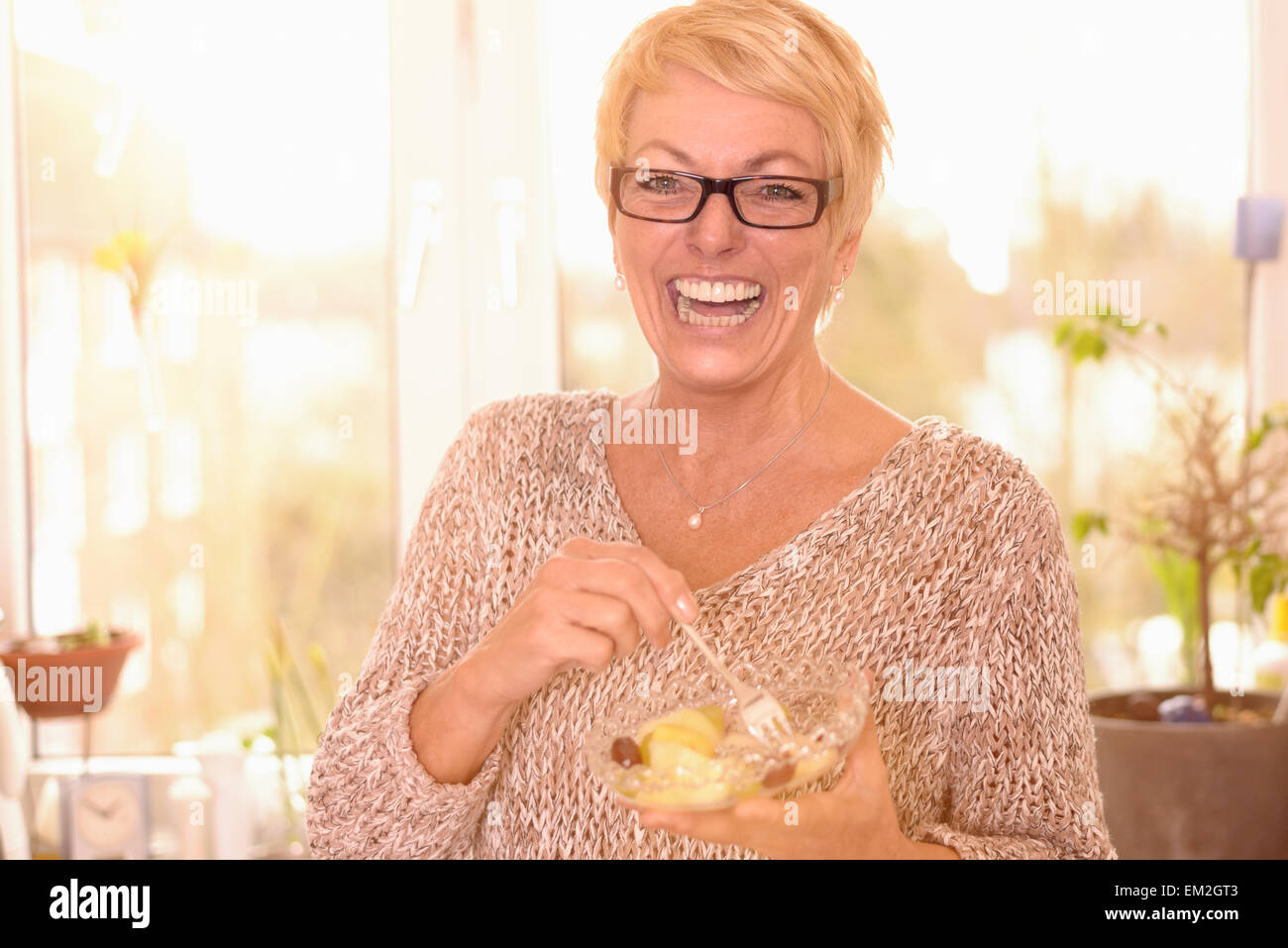 Vivace attraente donna di mezza età che indossano occhiali mangiare sano di una ciotola di insalata di frutta ricca di vitamine Foto Stock