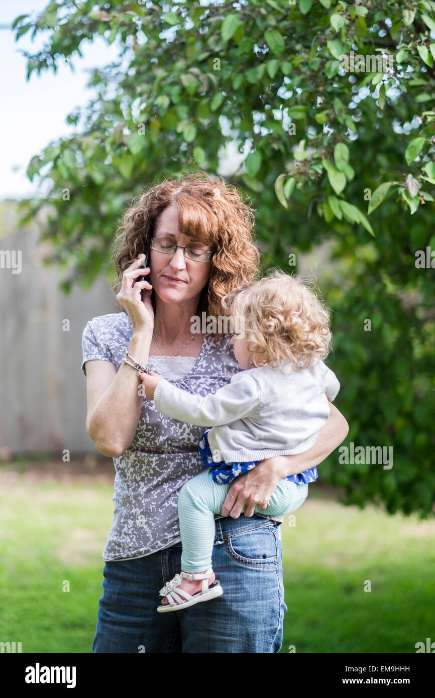 A 50 anno vecchia nonna tiene il suo bambino nipote sulla sua anca durante la ricezione di cattive notizie sul suo telefono cellulare all'esterno. Foto Stock