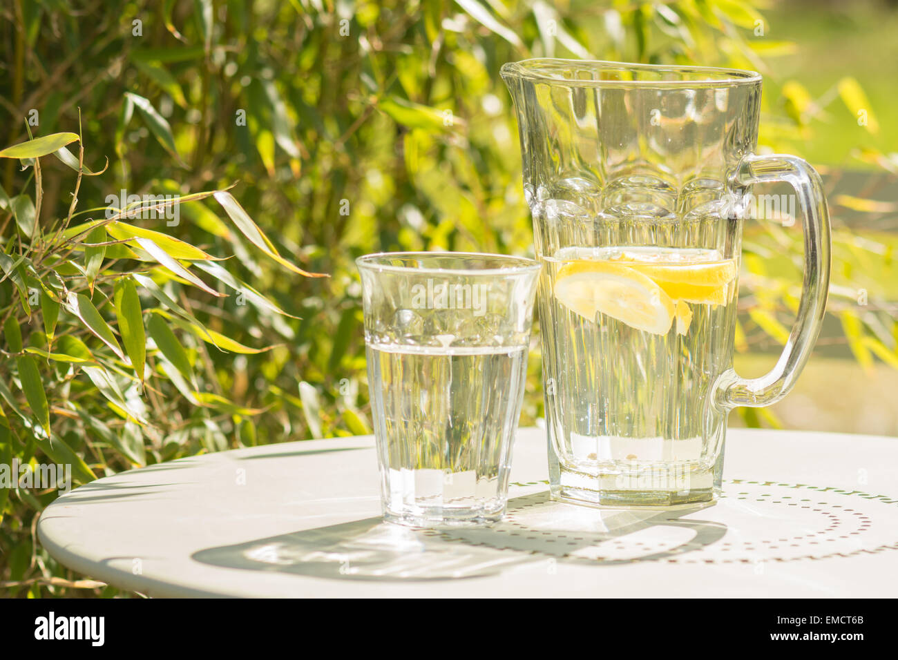 Estate - i riflessi del sole che splende attraverso la caraffa e bicchiere di acqua sul green bistro tavolo sulla terrazza giardino Foto Stock