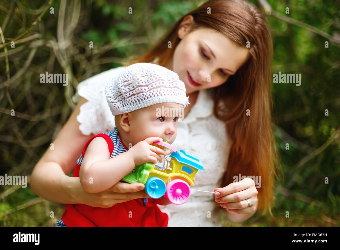 Carino il Toddler Baby ragazza seduta su Mom le mani, giocando con il giocattolo. Relax nel verde parco di estate. Messa a fuoco selettiva sul bambino agli occhi. Foto Stock