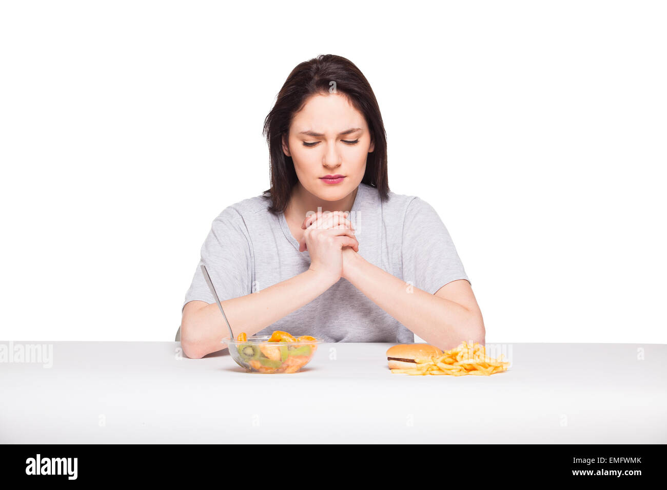 Una sana contro il cibo spazzatura concetto con un naturale donna ansante davanti pasto di frutta e patate fritte con hamburger, isolato su bianco Foto Stock
