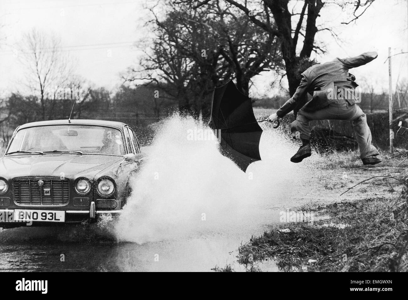 Un pedone con ombrello viene inzuppato come un automobile passa attraverso un enorme di una pozza d'acqua sul lato della strada in seguito a pesanti acquazzoni di pioggia in Oakington, Cambridgeshire. Il 5 maggio 1978. Foto Stock