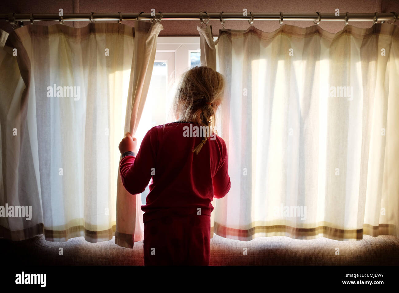 Una sonnolenta giovane ragazza in pigiama apre le tende a guardare fuori dalla finestra all'inizio di un nuovo giorno Foto Stock