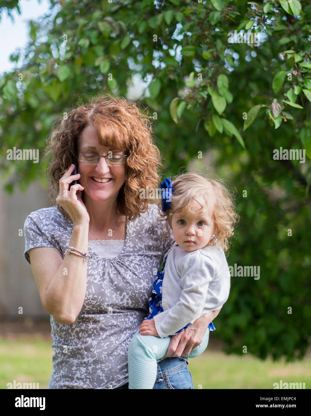 A 50 anno vecchia nonna tiene il suo bambino nipote sulla sua anca durante la conversazione sul suo telefono cellulare all'esterno. Stati Uniti d'America Foto Stock