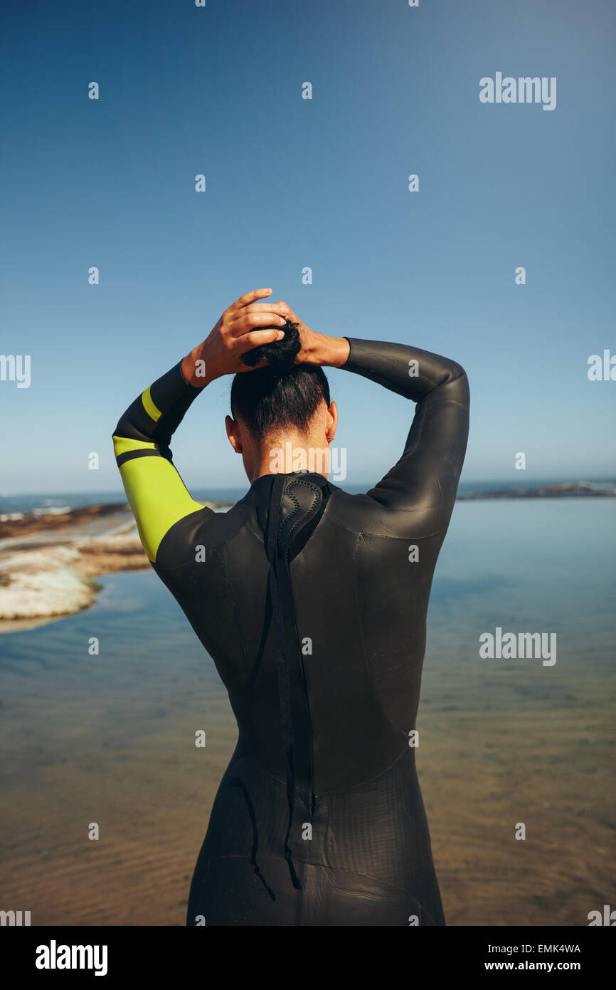 Vista posteriore del giovane triatleta sul lago della preparazione di una gara che indossa una muta. Triathlon femminile atleta legando i suoi capelli e ge Foto Stock