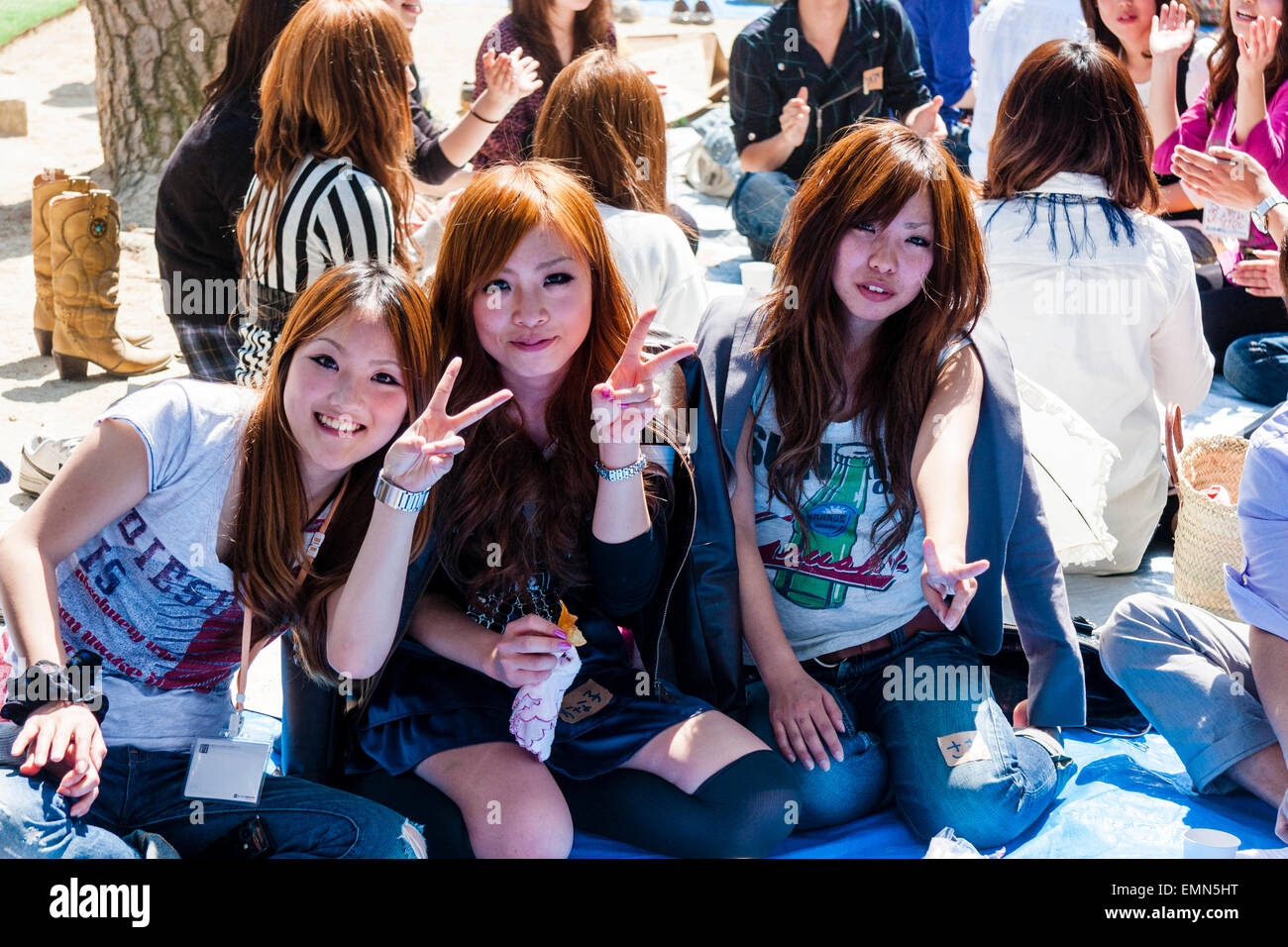 Tre giovani donne sedute con gli altri, guardando lo spettatore e dando ai due dita giapponese gesto di pace e sorridente durante la festa di fioritura dei ciliegi. Foto Stock