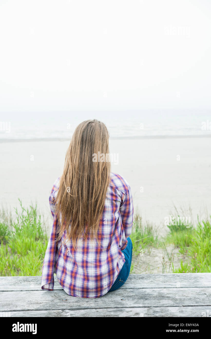 Vista posteriore della giovane donna con capelli lunghi biondi seduta sul vecchio picnic sulla spiaggia tabella rivolta verso l'oceano indossando plaid shirt Foto Stock