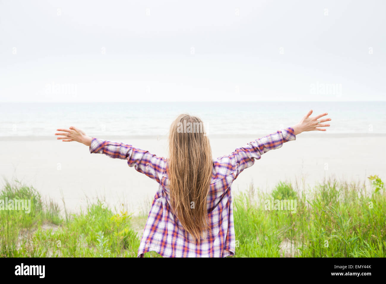 Vista posteriore della giovane donna con lunghi capelli biondi e le braccia tese sulla spiaggia vuota affacciata sull'oceano indossando plaid shirt Foto Stock