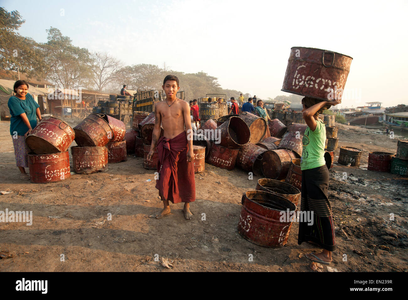 Un uomo birmano guardando la telecamera mentre si lavora sul Fiume Ayeyarwady circondato da sporco i fusti di olio e ragazza lavoratori Foto Stock