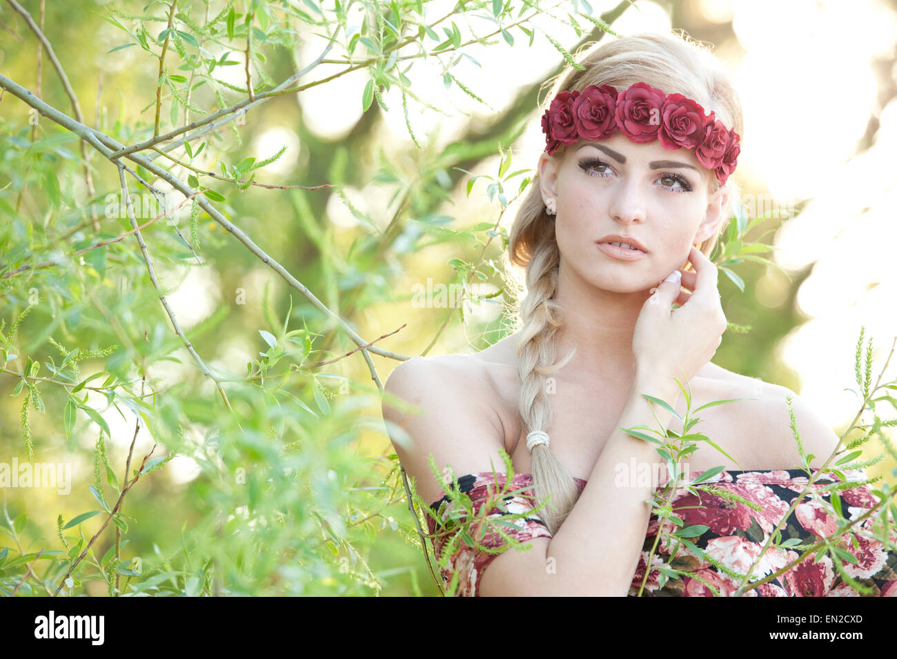 Un ritratto di una giovane splendida ragazza bionda cercando con la sua mano sulla guancia indossando un fiore rosso corona con un fiorito Foto Stock