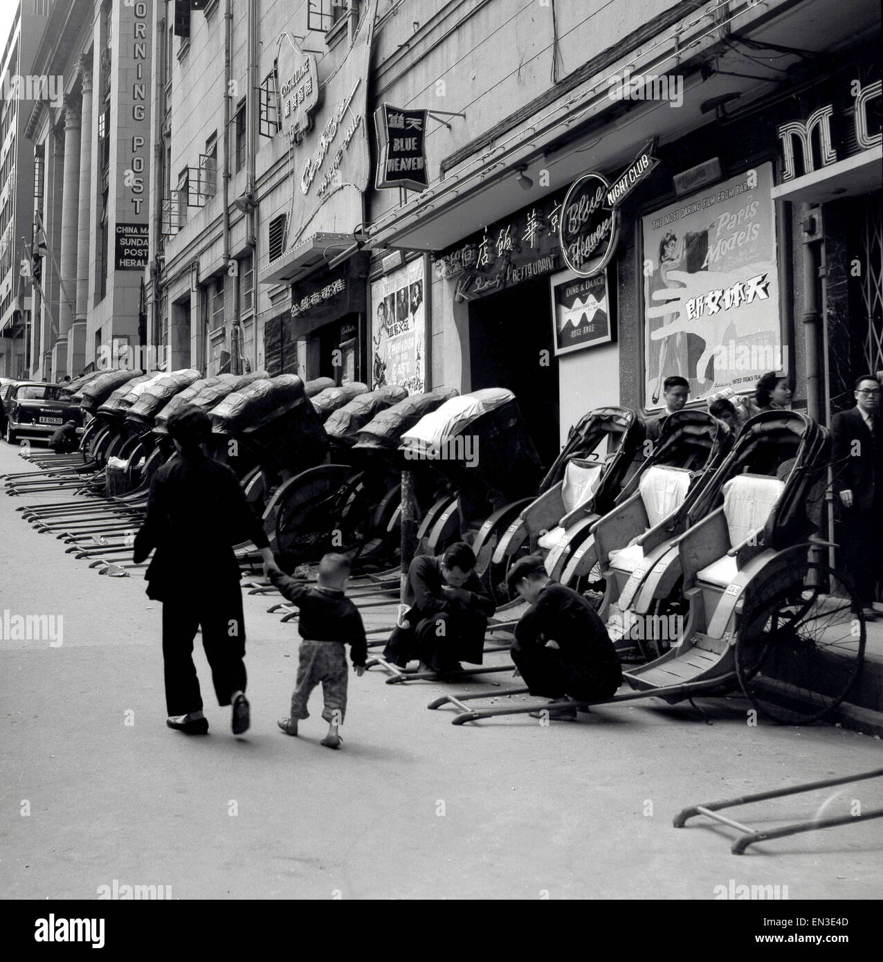 Foto storica degli anni '50, Hong Kong, su una strada laterale in pendenza, una fila di risciò tirati o carrelli passeggeri. La maggior parte dei risciò hanno le loro cappe in su, ma tre hanno le loro cappe giù che mostrano che sono disponibili per il noleggio. I loro operatori stanno giocando una partita per strada. I risciò sono stati una forma popolare di trasporto del taxi per i secoli, il loro nome proviene dalle japane per il trasporto umano-alimentato. Gli uffici del giornale della posta mattutina della Cina del sud possono essere visti ed i risciò sono parcheggiati fuori del teatro blu e del nightclub. Foto Stock