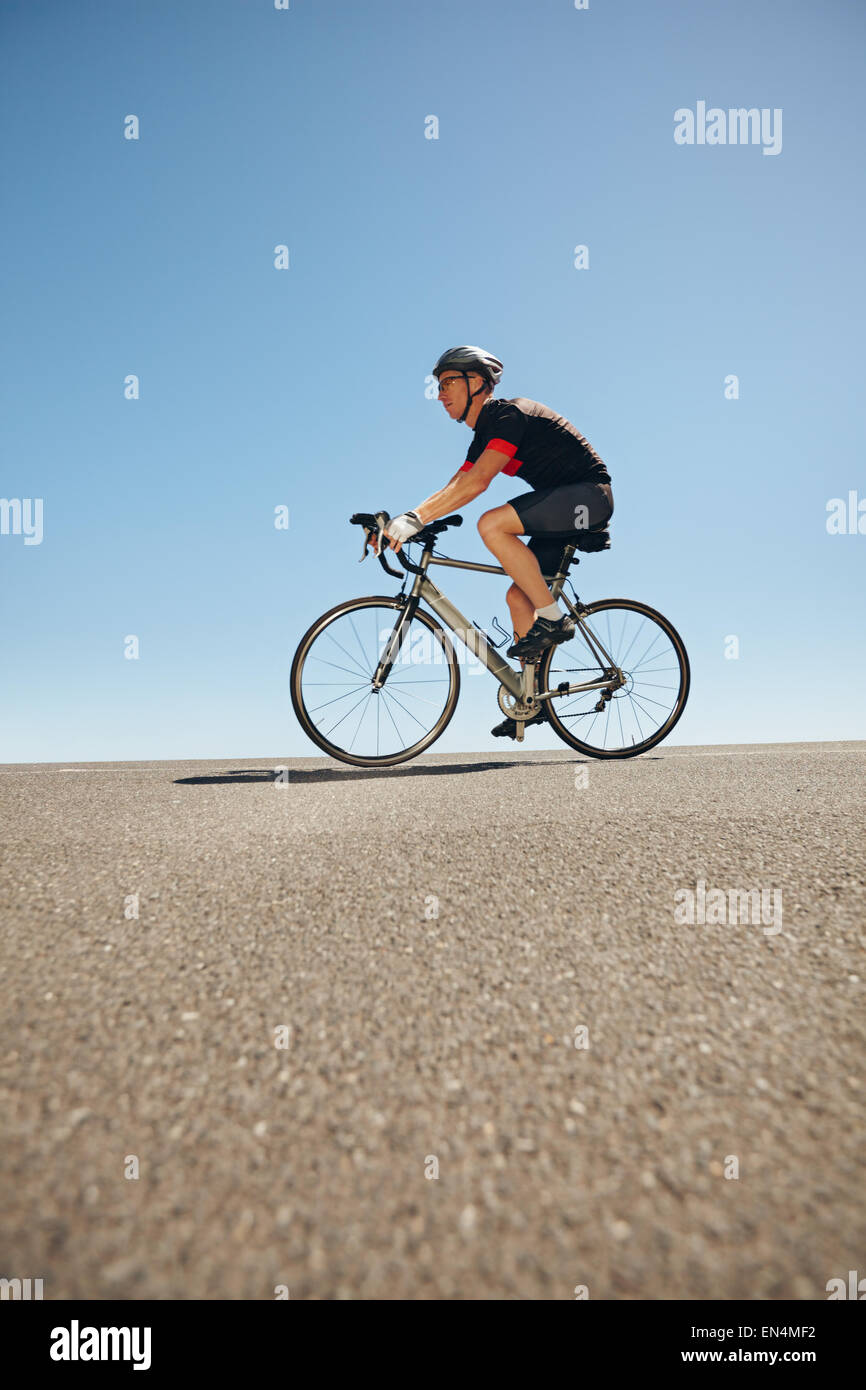 Vista laterale del ciclista maschio equitazione bicicletta su strada piana contro il cielo blu. Basso angolo vista del triatleta caucasica formazione per comp Foto Stock
