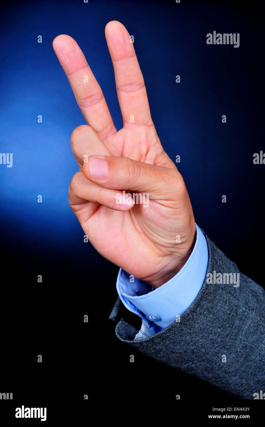 Primo piano della mano di un giovane imprenditore caucasico indossando un abito grigio dando un segno V su uno sfondo nero alleggerito in blu Foto Stock