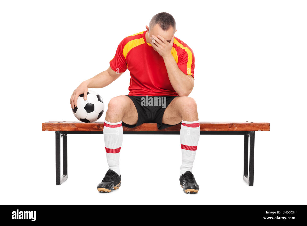 Preoccupato per il giocatore di football seduto su una panca in legno, tenendo una palla e nasconde la sua faccia con una sola mano isolati su sfondo bianco Foto Stock