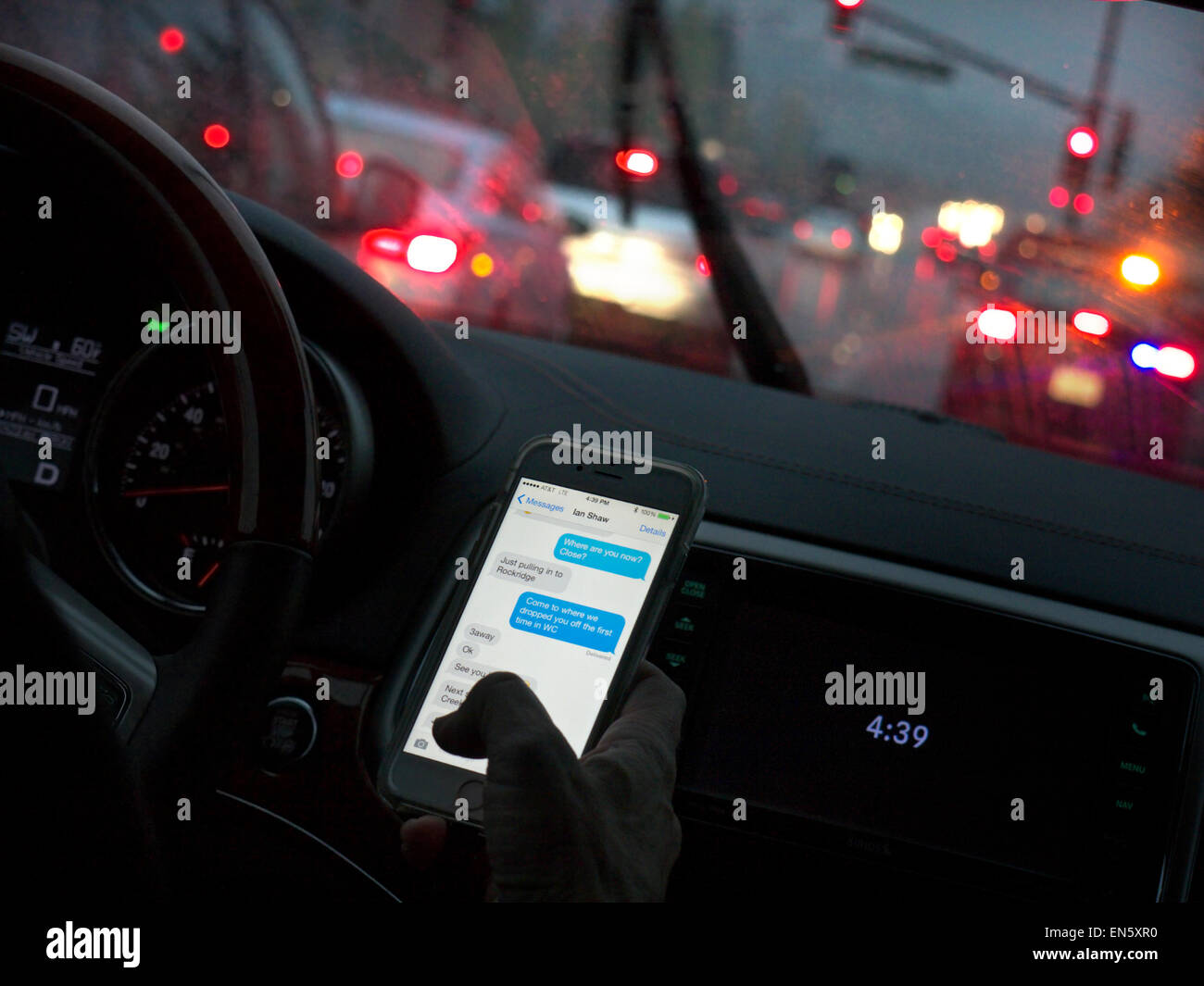 In auto driver femminile in l/h guidare il veicolo nascostamente texting con lo smartphone durante la guida su occupato bagnato dalla pioggia autostrada spazzata al crepuscolo California USA Foto Stock