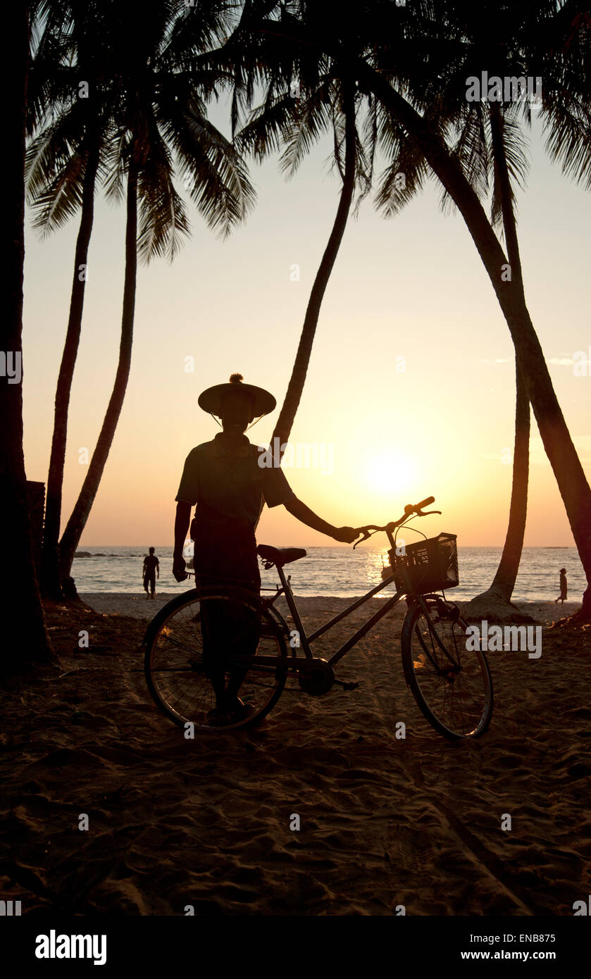 Silhouette di un uomo birmano indossando un cappello tradizionale in piedi con la sua bicicletta tra le palme sulla spiaggia di Ngapali Foto Stock