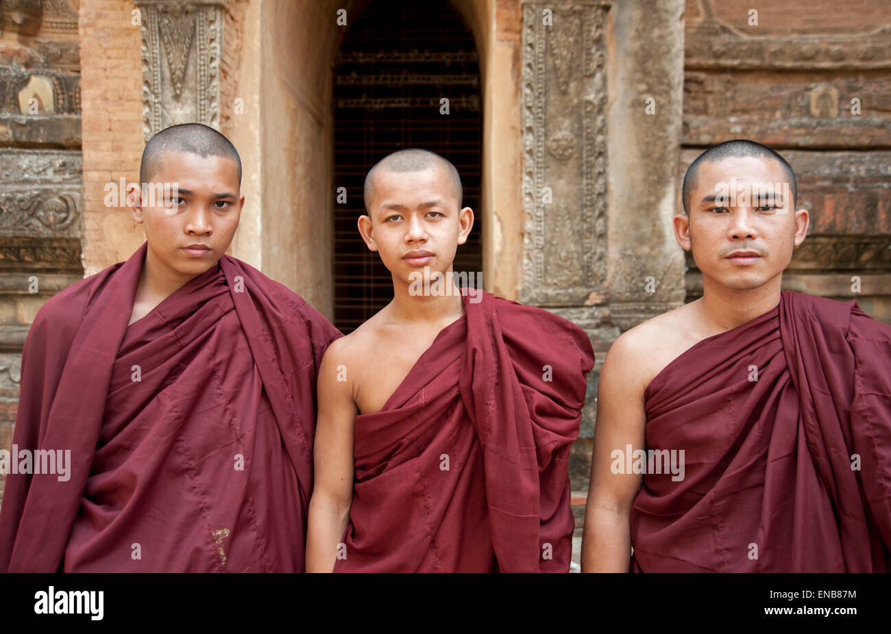 Ritratto di tre monaci buddisti che indossa vesti rosse contro il muro di pietra e ingresso di un tempio di Bagan MYANMAR Birmania Foto Stock
