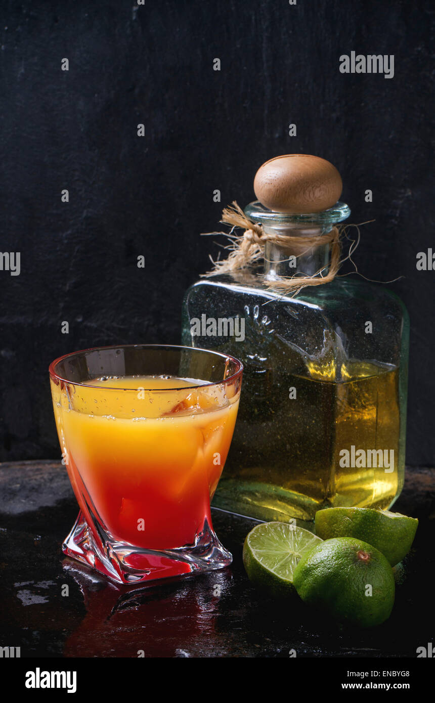 Bicchiere di tequila sunrise cocktail serviti con una bottiglia di tequila anejo affettato limes su sfondo nero. Rendendo la tequila sunris Foto Stock