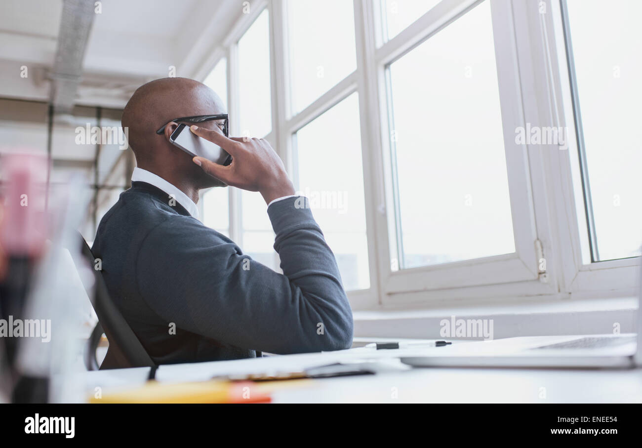 Giovane africano seduto alla sua scrivania parlando sul suo telefono cellulare in ufficio. African executive tramite telefono cellulare mentre si è al lavoro. Foto Stock