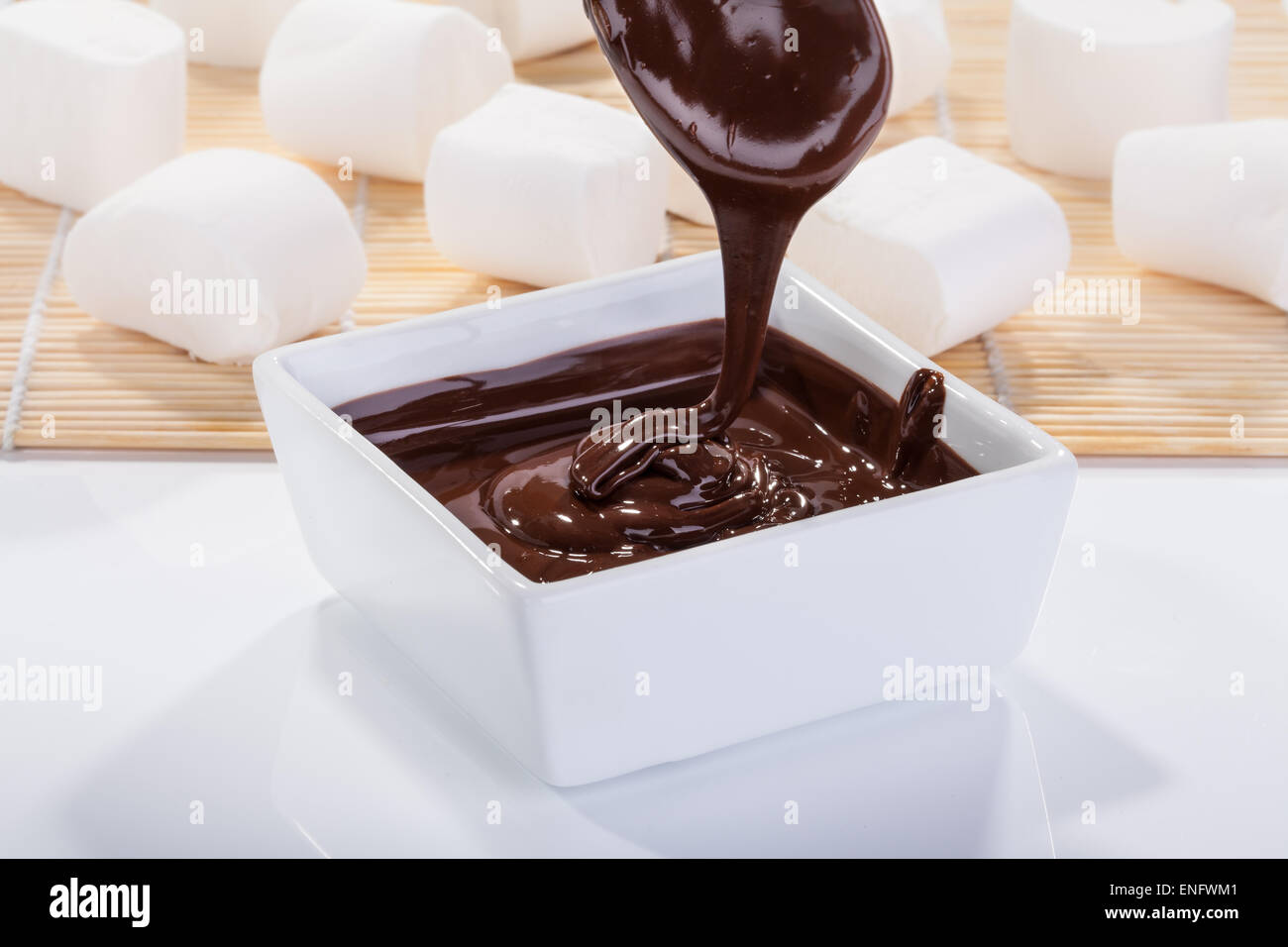 Salsa al cioccolato scorre verso il basso da un cucchiaio in una ciotola su un tavolo bianco Foto Stock