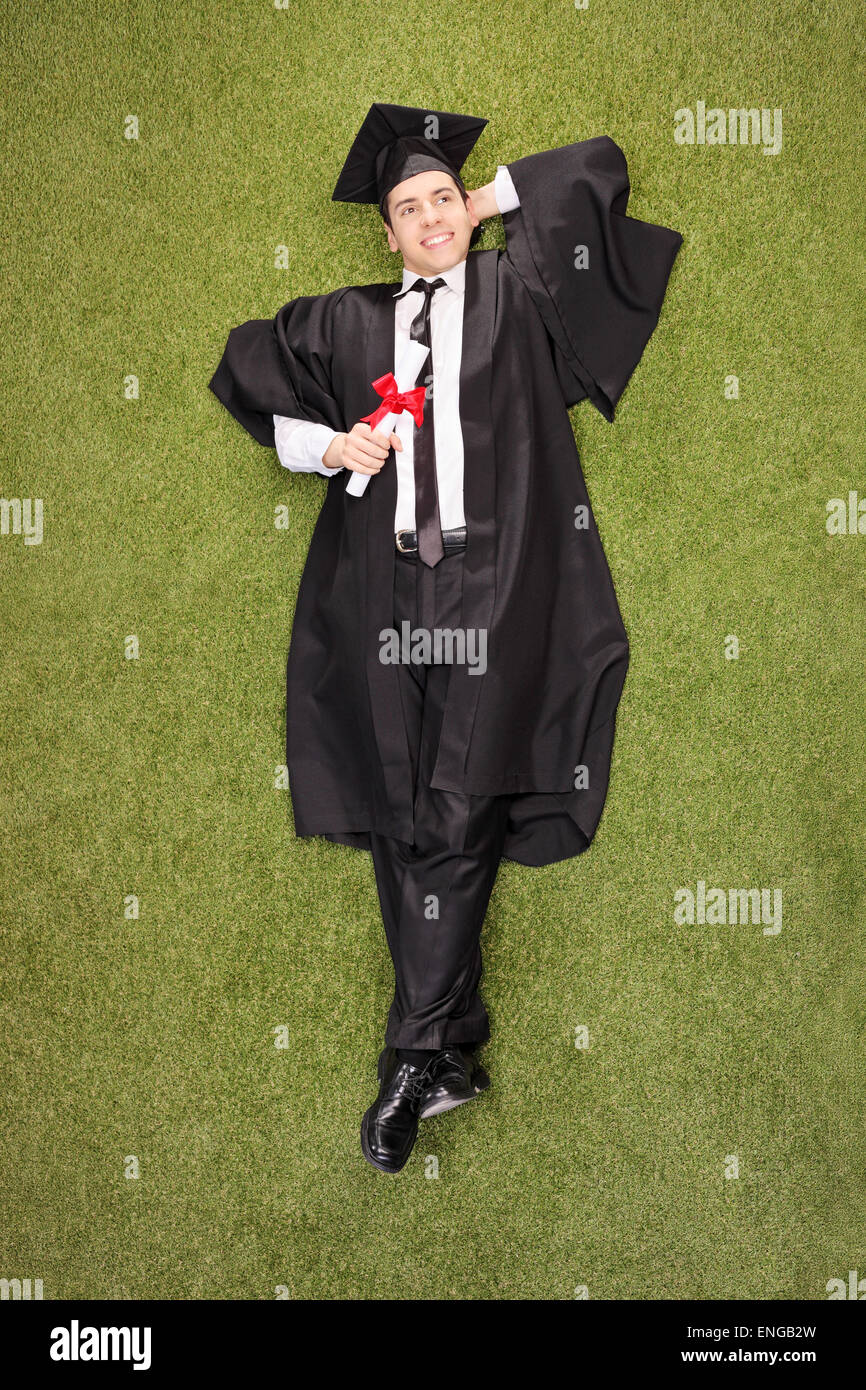 Colpo verticale di un pensieroso giovane college graduate in possesso di un diploma e giacente in un campo di erba Foto Stock