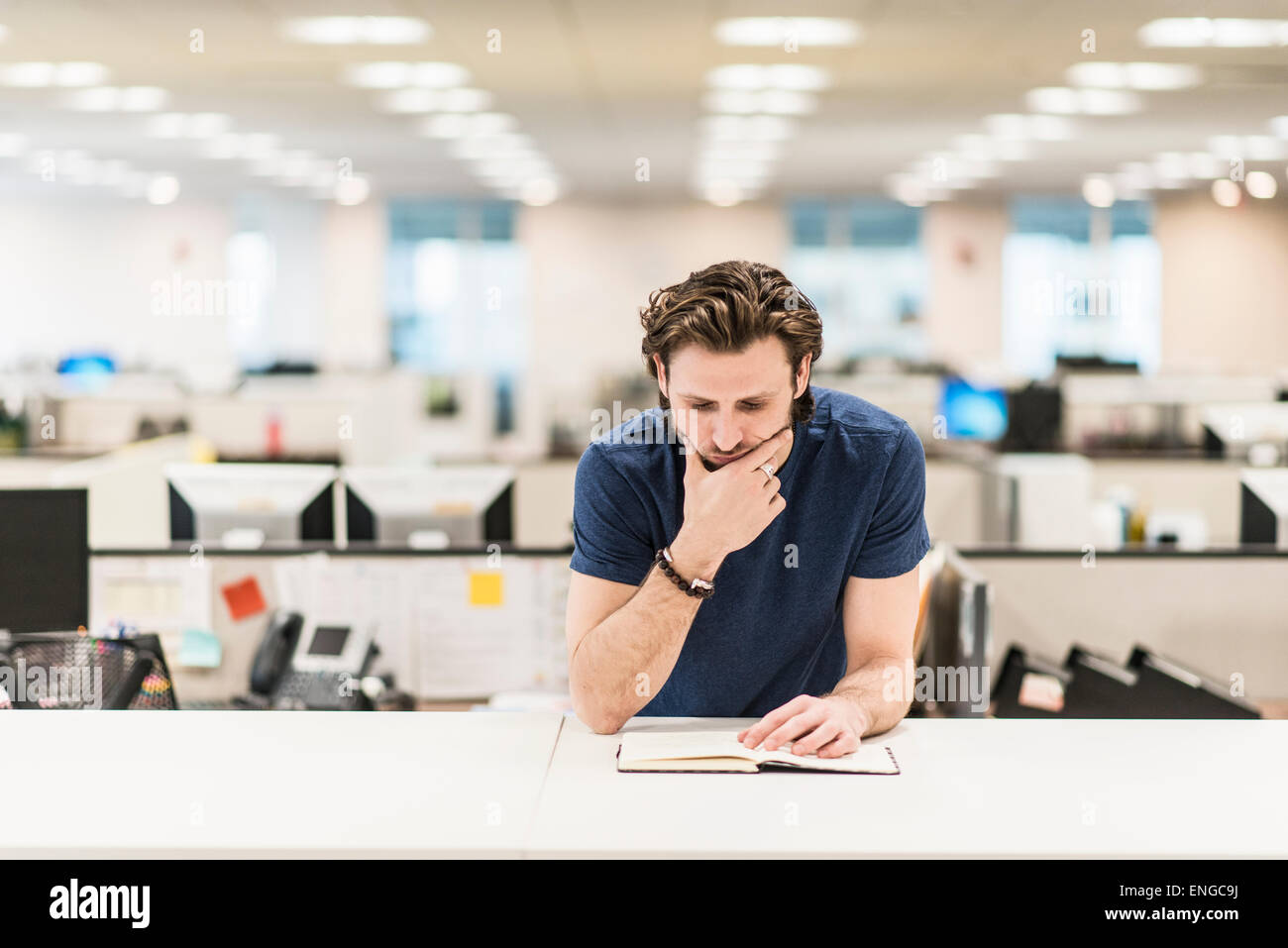 Un uomo appoggiato sul proprio gomito e guardando un libro aperto su una scrivania in ufficio. Foto Stock