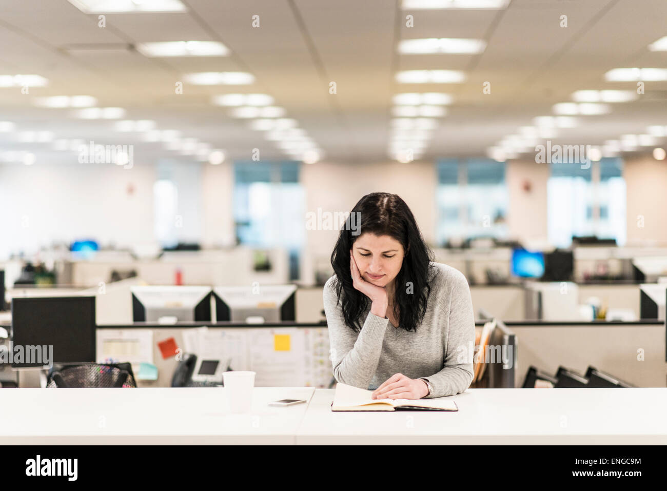 Una donna seduta con la sua mano appoggiata sul suo mento, la lettura di un libro a una scrivania. Foto Stock