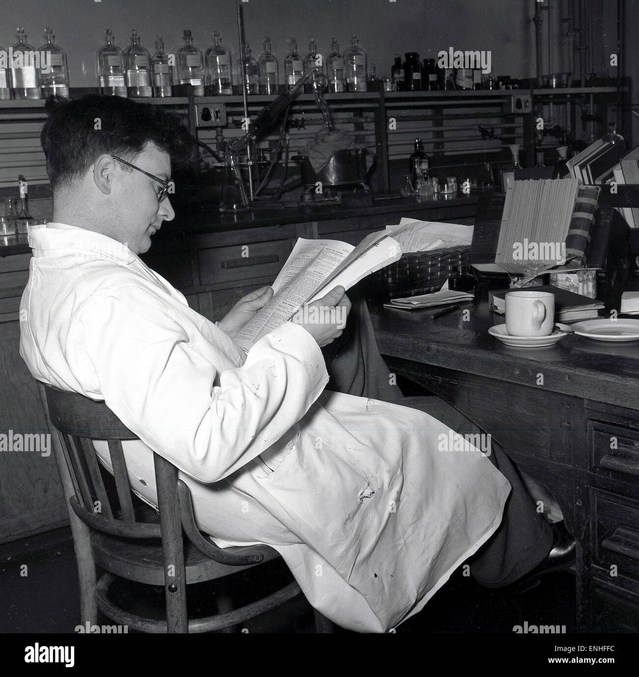 Degli anni Cinquanta, scienziato maschio bianco da indossare camice legge un ufficiale ad una scrivania in un medico di laboratorio di ricerca . Foto Stock