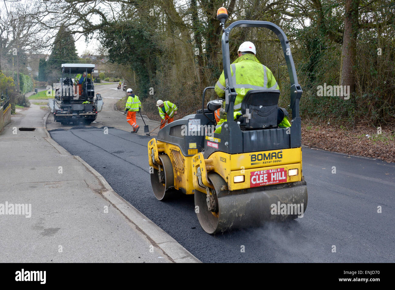 Uomini che lavorano in lavori stradali, riparazione e riparazione con rullo vibrante e macchina spalmatrice di asfalto indossando giacche ad alta visibilità e cappelli duri Essex UK Foto Stock