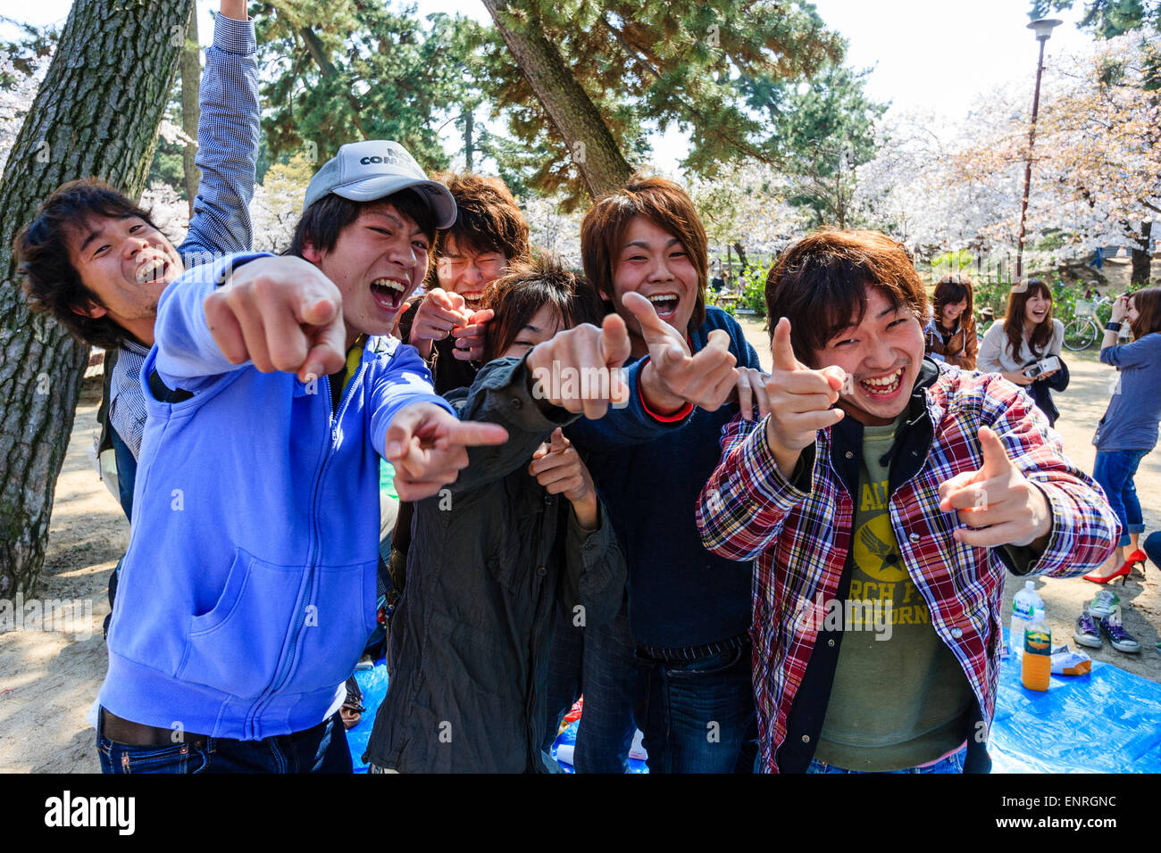 Gruppo di cinque adolescenti giapponesi amichevoli e una ragazza che si levano in piedi tutti sorridenti e che puntano allo spettatore durante il festival primaverile della fioritura dei ciliegi. Foto Stock