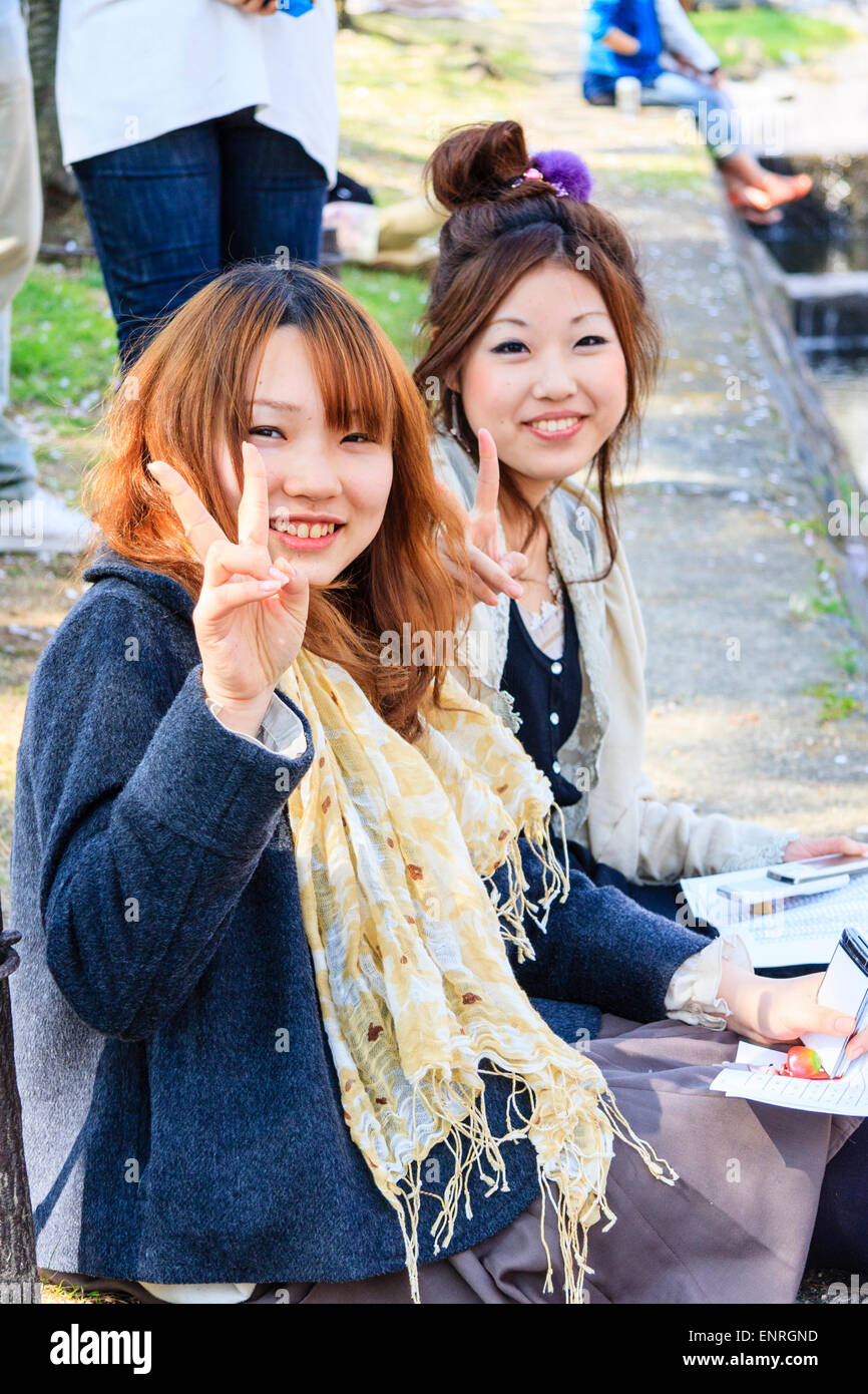 Due adolescenti giapponesi seduti sulla riva del fiume a Shukugawa in Giappone, entrambi si girarono per sorridere a spettatore e gesturando il segno di pace a due dita. Foto Stock
